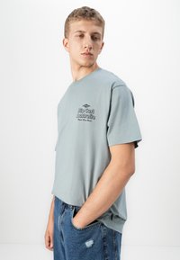 Young man wearing light blue Rip Curl Australia t-shirt and blue jeans with hand in pocket, looking downward to his left.