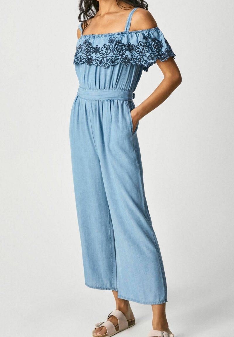 Jasnoniebieski dżinsowy kombinezon z odkrytymi ramionami, koronkowymi detalami przy dekolcie, z paskiem w talii oraz szerokimi nogawkami.