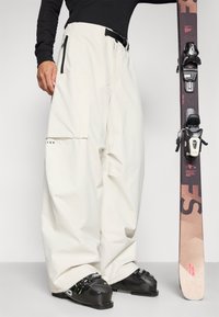 Bijele snowboard hlače s labavim krojem, s velikim bočnim džepovima i petljom za remen. Crne skijaške čizme i skija s vezovima drže se sa strane.