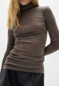 Hnědý dlouhý rukáv top s vysokým rolákem. Vyrobeno z texturované látky s přiléhavým střihem a jemným nařasením v pase.