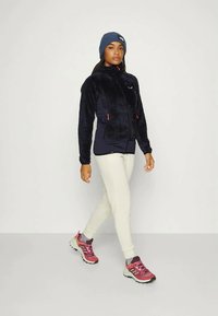 Naine kõnnib tumesinises fliisjakis, beežides pükstes, roosa ja punase värviga jooksutossudes ning sinises kudumispeas, lihtsa valge taustaga.