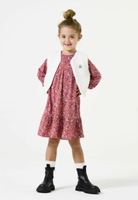 Rotes Kleid mit Paisleymuster und geraffter Taille, kombiniert mit einer weißen, strukturierten ärmellosen Weste. Schwarze Knöchelstiefel mit dicken Sohlen.