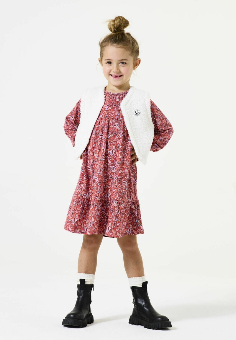 Rotes Kleid mit Paisleymuster und geraffter Taille, kombiniert mit einer weißen, strukturierten ärmellosen Weste. Schwarze Knöchelstiefel mit dicken Sohlen.