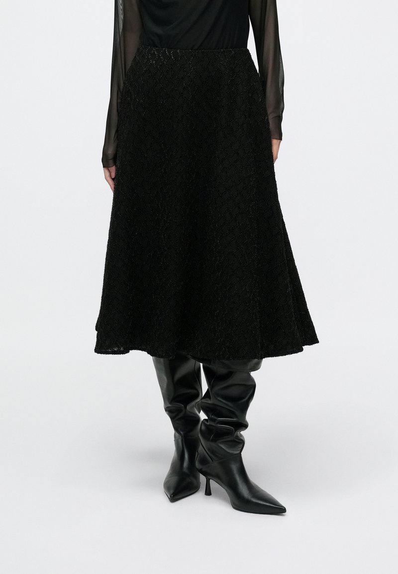 Sort tekstureret A-formet nederdel med et dekorativt diamantmønster, kombineret med elegante sorte knælange støvler og en gennemsigtig sort top.