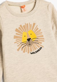 Sudadera de color beige claro con un gráfico de un león que tiene cara amarilla, acentos de melena marrones y la palabra "WILDNESS" en texto negro. Tejido suave y texturizado.