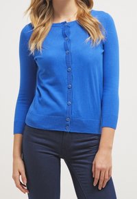 Blå stickad cardigan med rund halsringning, långa ärmar och knäppning; har en ribbad fåll och manschetter, slät textur.