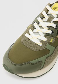 Sapatilha atlética verde com parte superior texturizada, detalhes em branco e amarelo, e atacadores redondos. A sola é leve e tem uma ligeira curvatura.