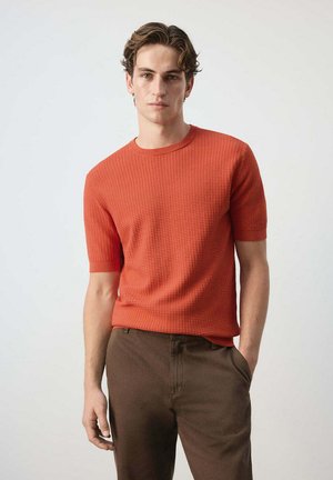 Junger Mann mit lockigem Haar trägt ein strukturiertes, orangefarbenes Kurzarmhemd und braune Hose, steht mit einer Hand in der Tasche.
