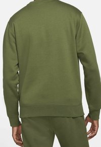 Bakifrån av en person i olivgrön långärmad sweatshirt och matchande byxor, stående med armarna avslappnat vid sidorna.