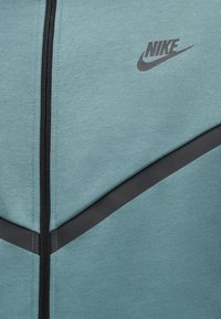 Υφασμάτινο σε ανοιχτό μπλε χρώμα με μαύρη διαγώνια ρίγα και φερμουάρ· λογότυπο Nike τυπωμένο μαύρο στην επάνω δεξιά πλευρά.