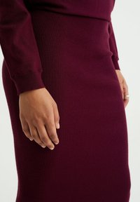 Vinröd ribbad stickad klänning med en figurnära silhuett. Har långa ärmar och strukturerad tyg. Närbild av en hand på klänningen.