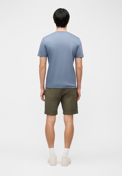 Άνδρας με μαύρα μαλλιά που φοράει ανοιχτό μπλε κοντομάνικη μπλούζα, ελαιοπράσινες κοντές βερμούδες με γυρισμένα μανίκια, λευκές κάλτσες και λευκά αθλητικά παπούτσια, στέκεται με την πλάτη προς τα έξω.