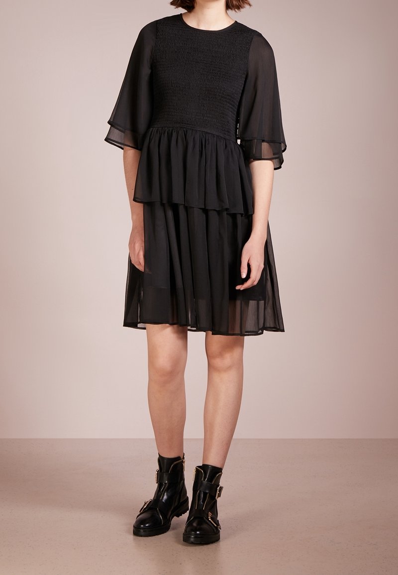 Femme portant une robe noire superposée avec des manches courtes transparentes et des bottines noires à boucles sur un fond neutre.