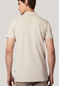 Jasno beżowa koszulka polo z krótkim rękawem, miękka tkanina, gładka tekstura i mały detal z logo na tyle kołnierzyka.