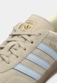 Μπεζ δερμάτινο αθλητικό παπούτσι με τρεις μπλε ρίγες, χρυσό λογότυπο στη γλώσσα και κορδόνια ανοιχτού χρώματος. Διαθέτει υφή στη σόλα.