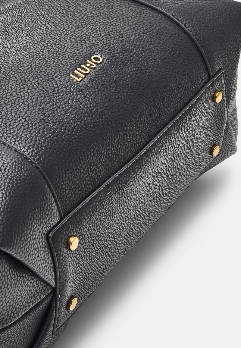 Sac en cuir noir avec une surface texturée, du matériel en or et un logo bien en vue. Les accents comprennent des clous dorés sur les côtés.
