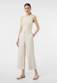 Kremowa bluzka bez rękawów z teksturowanym dekoltem, zestawiona z białymi spodniami o wysokim stanie i szerokich nogawkach. Noszone z naturalnymi, plecionymi klapkami.