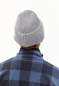 Grijze gebreide muts met een geribbelde textuur, met een taps toelopende bovenkant. Draagt bij een blauw-zwarte geruite fleecejas.