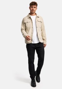 Beige Knopfjacket mit zwei Brusttaschen, getragen über einem weißen T-Shirt. Kombiniert mit schwarzen Hosen und schwarzen Schuhen, die lässigen Stil präsentieren.