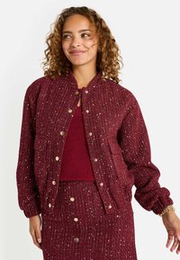 Chaqueta bomber acolchada burdeos con acentos de hilo metálico, botones a presión y cuello redondeado. Tejido texturizado con puños elásticos.