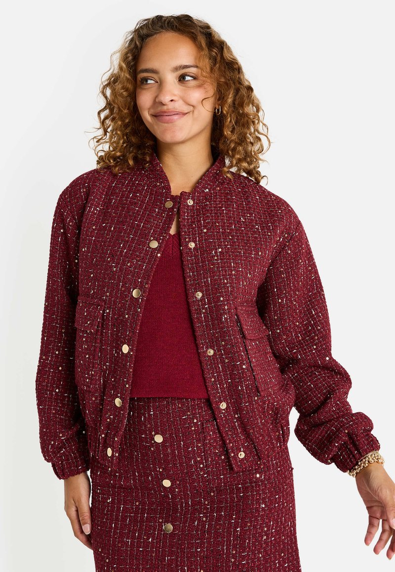 Chaqueta bomber acolchada burdeos con acentos de hilo metálico, botones a presión y cuello redondeado. Tejido texturizado con puños elásticos.