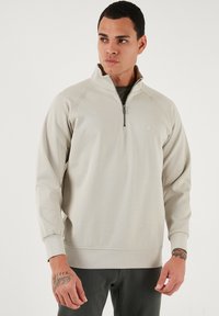 Svijetlosmeđi dukserica na zip s visokim ovratnikom, raglan rukavima i rebrastim manžetama, s malim logotipom na prsima. Glatka tekstura.