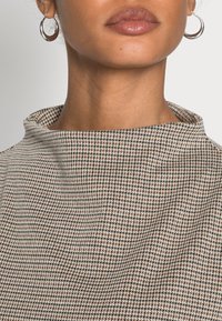 Tissu à motif pied-de-poule marron et blanc avec un col montant et une surface texturée. Des boucles d'oreilles argentées en forme de créole ajoutent un détail subtil.