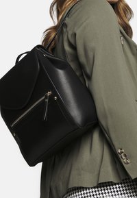 Sac à dos en cuir noir avec des accessoires en or, poche zippée à l'avant, rabat supérieur et poignée courte ; porté sur l'épaule avec une veste vert olive.