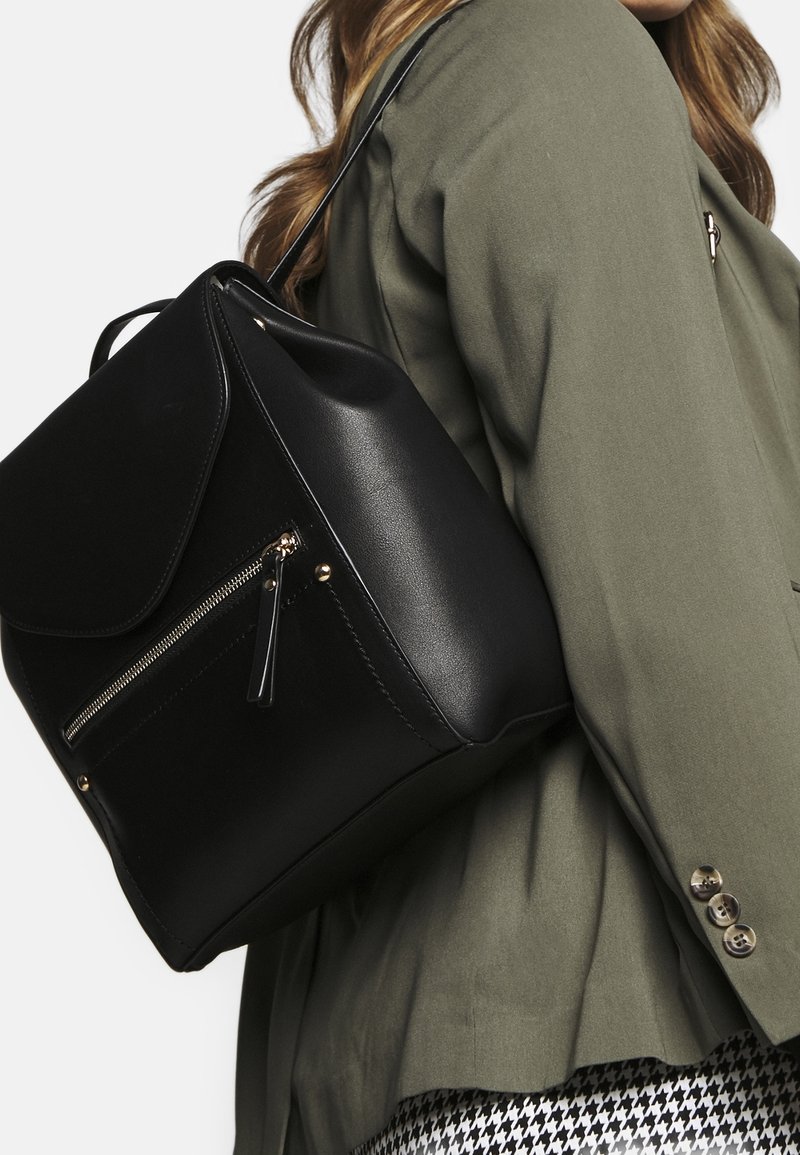 Sac à dos en cuir noir avec des accessoires en or, poche zippée à l'avant, rabat supérieur et poignée courte ; porté sur l'épaule avec une veste vert olive.