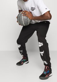 Svarta träningsbyxor med laglogotyper och grafik, kombinerat med en vit T-shirt som har en cirkulär logotyp och färgglada sportskor.