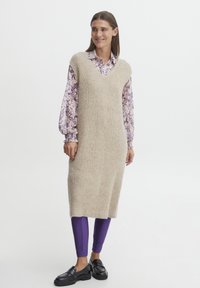 Abito senza maniche a coste beige con scollo a V, sovrapposto a una blusa floreale viola e abbinato a leggings viola e mocassini neri.