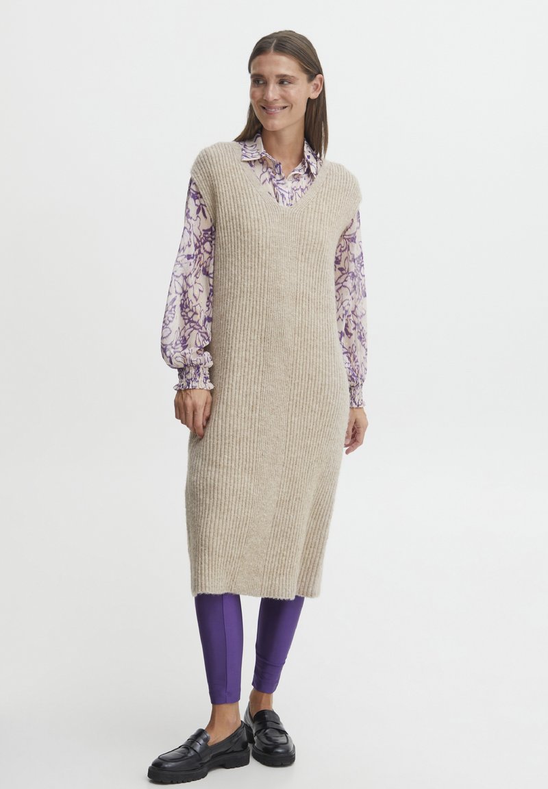 Abito senza maniche a coste beige con scollo a V, sovrapposto a una blusa floreale viola e abbinato a leggings viola e mocassini neri.