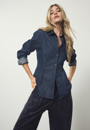 Dunkelblaue Denim-Hemd mit einem spitzen Kragen, langen Ärmeln und diagonal verlaufenden Nähten, kombiniert mit lockeren navyblauen Cordhosen.