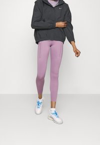 Světlé fialové legíny s bočními kapsami na zip ve spojení s tmavě šedou mikinou s kapucí. Bílá teniska s modrými akcenty doplňuje outfit.