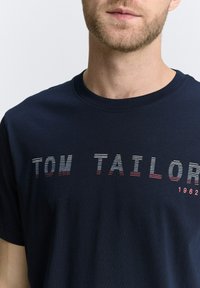 Camiseta de algodón azul marino con cuello redondo, que presenta un texto en rayas horizontales "TOM TAILOR" en blanco y rojo, con un pequeño detalle del año "1962".