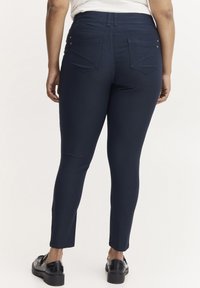 Marineblaue Skinny-Jeans mit glatter Textur, mittelhoher Taille und zwei Gesäßtaschen. Mit silberfarbenen Akzenten für zusätzlichen Detailreichtum.