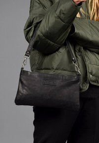 Bolso cruzado de piel negra con una textura suave, forma rectangular, correa ajustable y herrajes metálicos. Exhibido sobre un fondo gris.