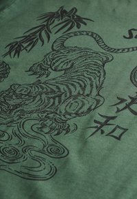 Groene stof met een zwart grafisch ontwerp van een tijger te midden van draaiende patronen en gestileerde tekst, waarmee complexe lijnen en texturen worden weergegeven.