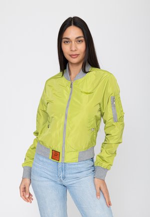 Femme aux longs cheveux foncés portant une veste bomber vert clair avec des poignets gris et un jean bleu clair, debout devant un fond uni.
