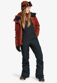 Chaqueta de nieve roja y negra con capucha forrada de piel, combinada con un overol negro. Modelo lleva un gorro de lana marrón y gafas de esquí. Guantes negros completan el look.