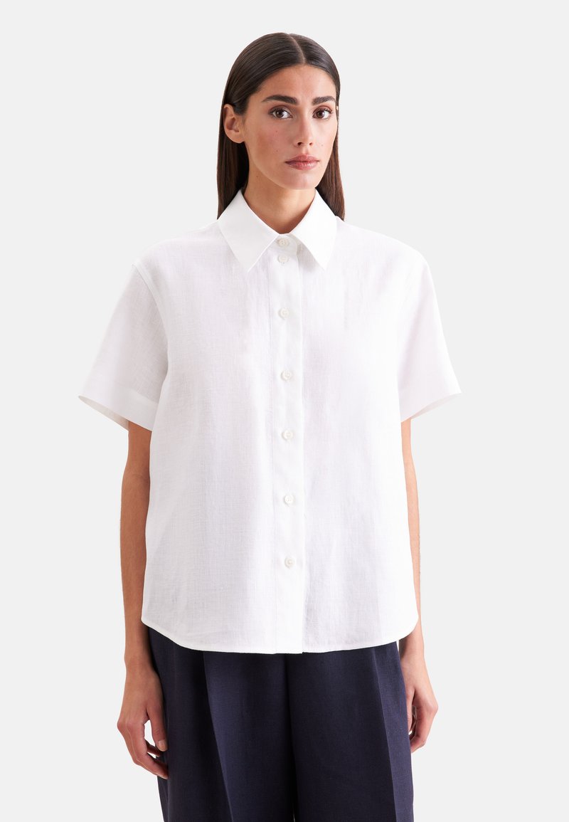 Femme aux cheveux raides et foncés porte une chemise blanche à manches courtes boutonnée et un pantalon ample foncé sur un fond uni.