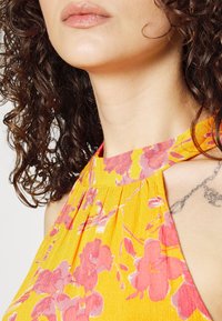 Žena s kudrnatými tmavými vlasy, oblečená ve žlutých šatech s holými rameny a růžovými květinovými vzory, s částečným pohledem na jemný řetízkový tetování na jejím rameni.