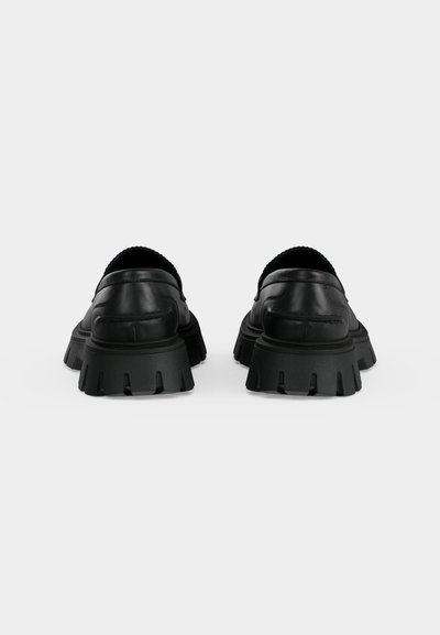 Paire de mocassins en cuir noir avec semelles en caoutchouc épaisses et volumineuses, présentés de dos sur un fond clair uni.