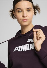 Felpa viola con un grande logo bianco "PUMA", dotata di un cordino e una texture morbida. In primo piano, una persona tiene il cordino.