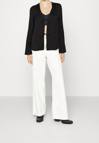 Chemise noire à manches longues, à boutons, avec décolleté en V, détail de nœud sur le devant et coupe décontractée, associée à un pantalon large blanc et des bottines noires.