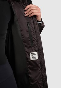 Dunkler, isolierter Mantel mit einer glatten, glänzenden Außenseite und einer Reißverschlusstasche innen. Ein Etikett trägt die Aufschrift: „Eine warme Umarmung aus Stoff.“