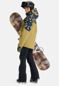 Snowboardzista nosi żółtą kurtkę z niebieskim wzorzystym kapturem, czarne spodnie i rękawice. Za nim znajduje się wielokolorowa deska snowboardowa.