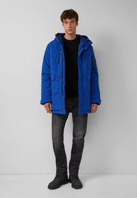 Blauer isolierter Parka mit Kapuze, ausgestattet mit Reißverschlusstaschen und einem Knopfverschluss, getragen über einem schwarzen Pullover und grauen Jeans.