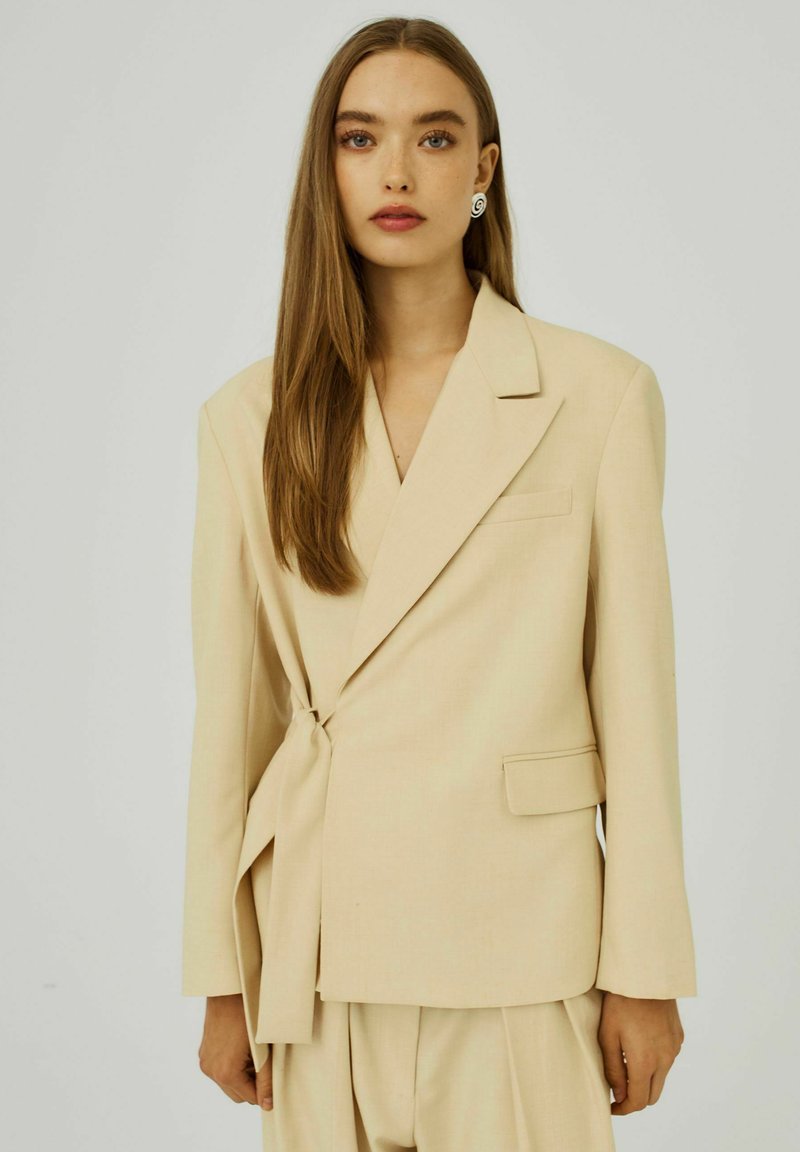 Beige zavinovací blazer s dlouhými rukávy, jednou kapsou a bočním vázáním, vyrobený z hladkého materiálu s jemnou strukturou.