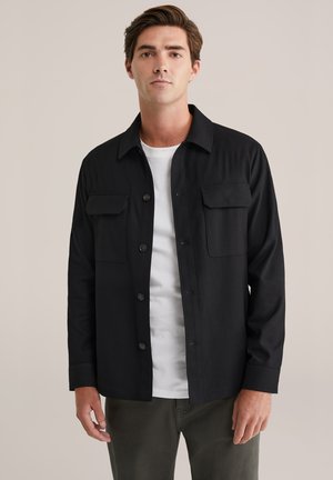 Camicia nera con bottoni in finitura opaca, dotata di due tasche sul petto, maniche lunghe e collo classico. Indossata sopra una t-shirt bianca.